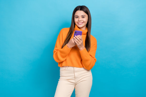 照片中，满意的可爱女孩直发，穿着橙色毛衣，拿着智能手机，在蓝色背景下阅读帖子图片下载