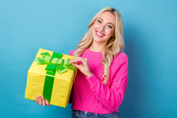 照片开朗愉快可爱的女孩与卷发穿着针织毛衣抱着礼物盒孤立的蓝色背景图片下载