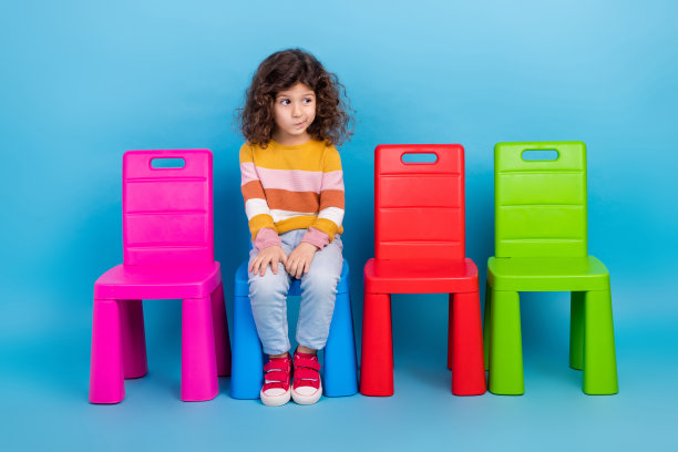 小女孩的全身照片，穿着时髦的休闲装条纹毛衣，坐在幼儿园的椅子上，在蓝色的背景上显得孤立图片下载