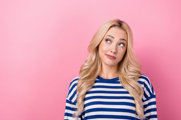 照片中的年轻梦幻可爱的女人穿着条纹衬衫，金发卷曲的头发，看起来空白的空间，选择化妆品孤立在粉红色的背景图片下载