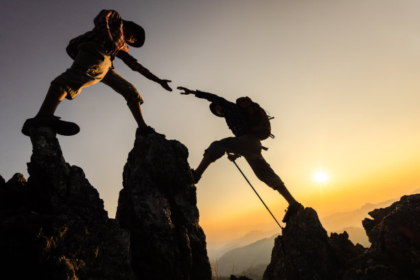 日出时山谷上方的岩石山脊上，两名登山者伸出援助之手。团队合作，团结工作。图片下载