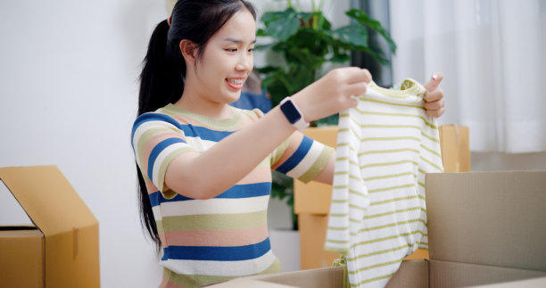 微笑的年轻女子从盒子里拿出一件衬衫，拍了一张照片。图片下载