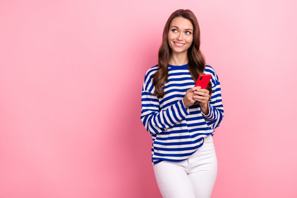 照片中开朗可爱的女人卷发，穿着条纹衬衫，拿着智能手机，在粉红色的背景下看起来空无一物图片下载