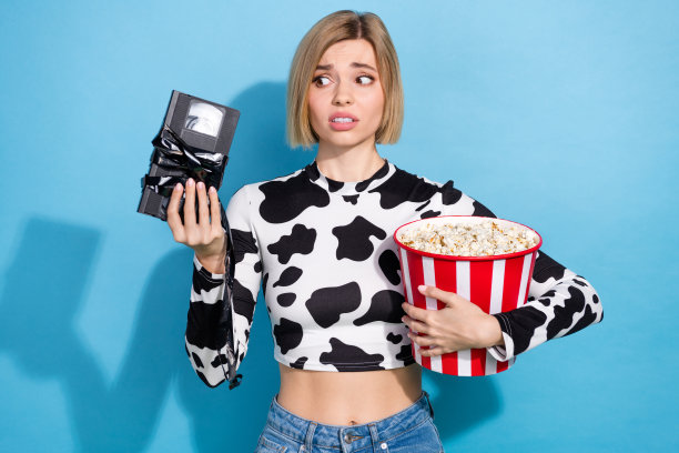 年轻女子的照片鲍勃头发穿斑点狗印花短上衣拿电影磁带困惑如何看电影吃爆米花孤立在蓝色的背景图片下载