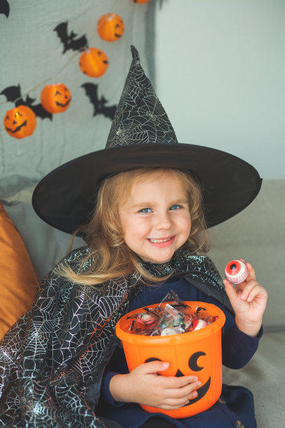 可爱的小女孩3岁，穿着女巫服装，拿着装满糖果的南瓜桶在家里庆祝万圣节。图片下载
