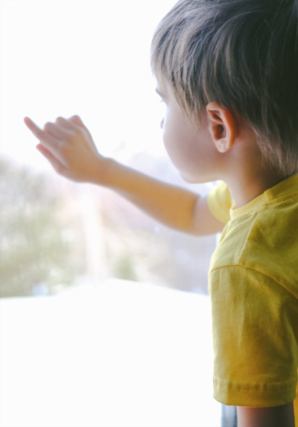 特写:可爱的小男孩用手指在窗玻璃上画画或写字，可爱的孩子坐在窗边等人回家，孩子手绘的背影图片下载