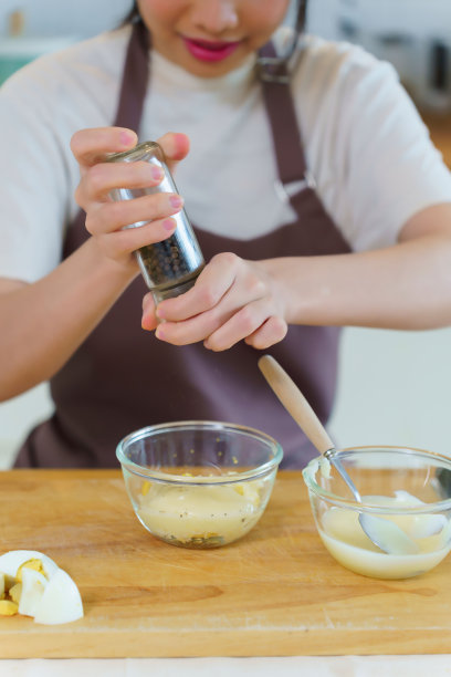 亚洲女性在家里的厨房里，一边准备早餐的食材，一边将蛋黄酱和胡椒粉加入到拌好的沙拉酱中图片下载
