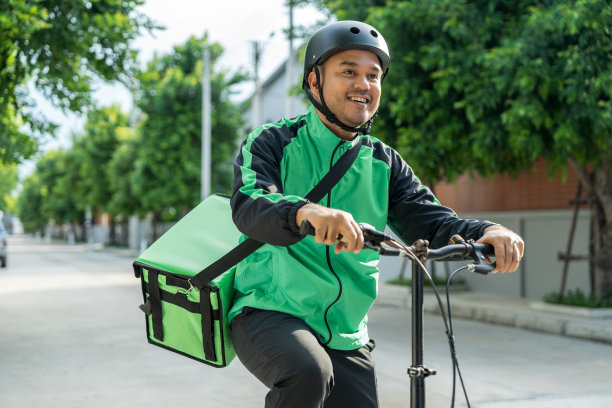 肖像骑手送餐员穿着绿色制服戴着头盔骑着自行车送餐给顾客。快乐快递员用绿色背包将货物运送给客户。图片下载