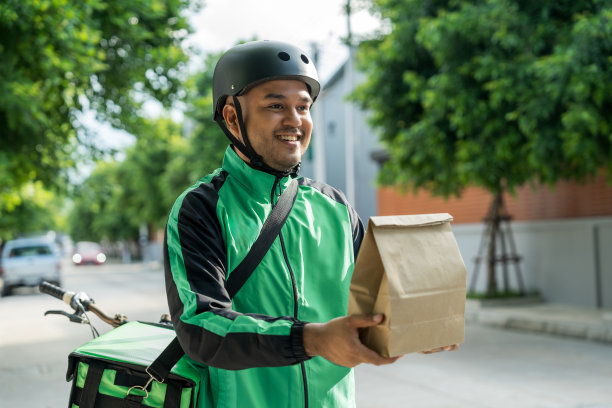 肖像骑手送餐员穿着绿色制服戴着头盔骑着自行车送餐给顾客。快乐快递员用绿色背包将货物运送给客户。图片下载