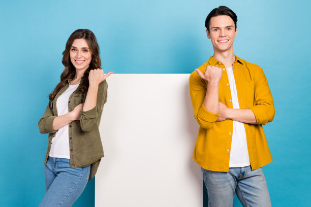 照片中两个年轻人穿着时尚随意，直视手指，纸广告牌，促销广告，空白空间孤立在蓝色背景上图片下载