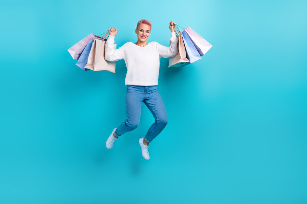 跳起来粗心的女孩的全身长度的照片，双手拿着新衣服购物季广告的袋子孤立在蓝色背景图片下载