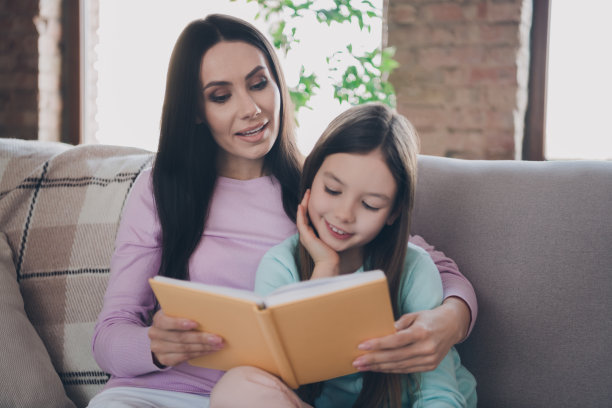 梦幻闪亮的母亲女儿穿着休闲装坐在沙发上享受有趣的书在室内房间的照片图片下载