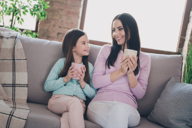 两个有趣的女孩的照片小女儿年轻的妈妈坐在舒适的沙发拿热温暖的饮料杯饮料家平坦的房子图片下载