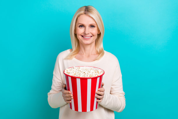 照片中漂亮开朗的女人穿着白衬衫，拿着大的爆米花桶，孤立的绿松石色背景图片下载