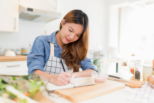 年轻的亚洲女性在笔记本上写食物食谱，一边吃酸奶，一边准备新鲜蔬菜沙拉，在家用健康的生活方式在现代厨房里做健康的早餐图片下载