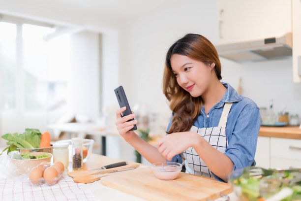 年轻的亚洲女性一边吃酸奶，一边用智能手机搜索食物食谱，一边准备新鲜蔬菜沙拉，在家用健康的生活方式在现代厨房里烹饪健康的早餐图片下载