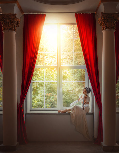 艺术照片真人。幻想女小提琴手演奏古典古典风格的小提琴。女孩坐在窗台上，花园的阳光闪闪发光。性感美女脸白色象牙色长丝质复古长裙图片下载