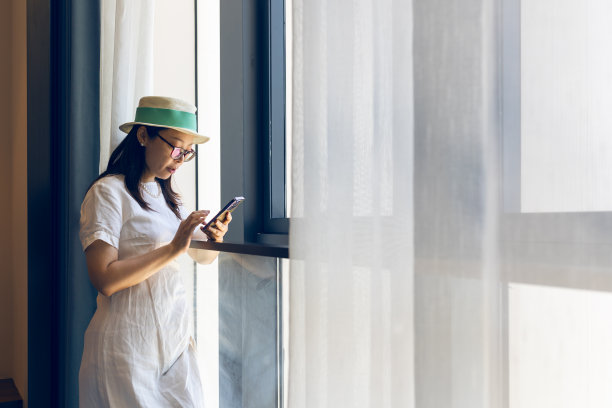 年轻的亚洲女子站在酒店窗口，用手机观看城市的天际线图片下载