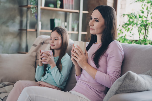 甜美可爱的女儿妈妈穿着休闲装在公寓房间里看电视喝可可微笑的照片图片下载