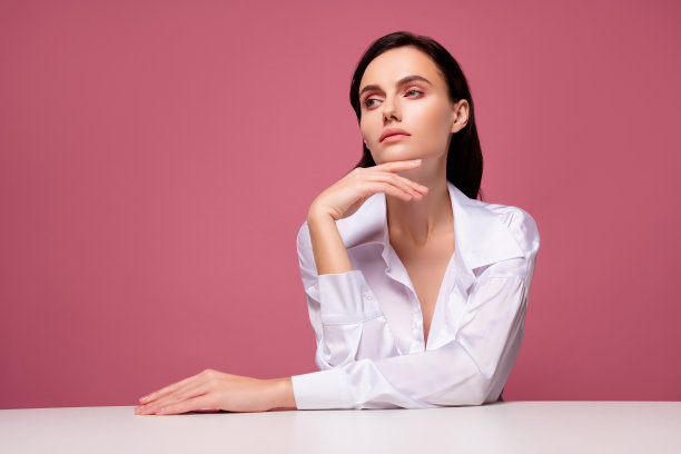 照片中的女孩坐在桌子上，摸着下巴，手看着优雅的粉红色空白空间，背景孤立图片下载