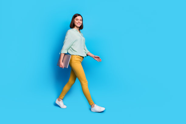 成功女性的全身照片，穿着青色衬衫，黄色裤子，在蓝色背景上孤立的笔记本电脑图片下载