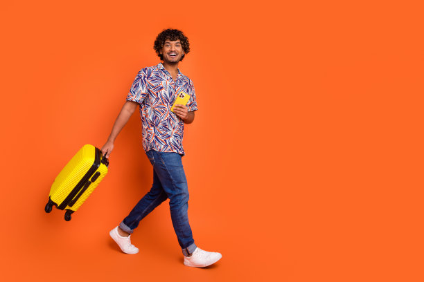 英俊的年轻人走路的全身照片，手持手提箱装置，穿着时尚的彩色服装，孤立在橙色背景上图片下载