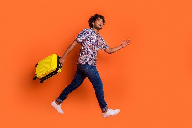 有魅力的年轻人抱着行李跑着的全身照片，他穿着时髦的彩色衣服，孤立在橙色的背景上图片下载