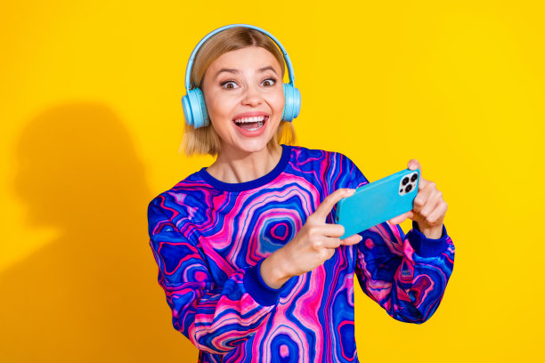 漂亮的年轻女孩的照片肖像，耳机玩兴奋的手机游戏，穿着时髦的彩色蓝色服装孤立在黄色的背景图片下载