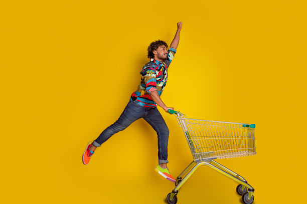 漂亮的年轻人的全尺寸照片推着超市推车飞行超人穿着时髦的彩色服装孤立的黄色背景图片下载