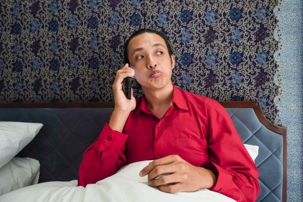 一头长发，身穿红色长袖衬衫的年轻亚洲男子对自己在家或酒店远程办公时在智能手机上大喊大叫感到沮丧和愤怒。自由职业者的概念。图片下载