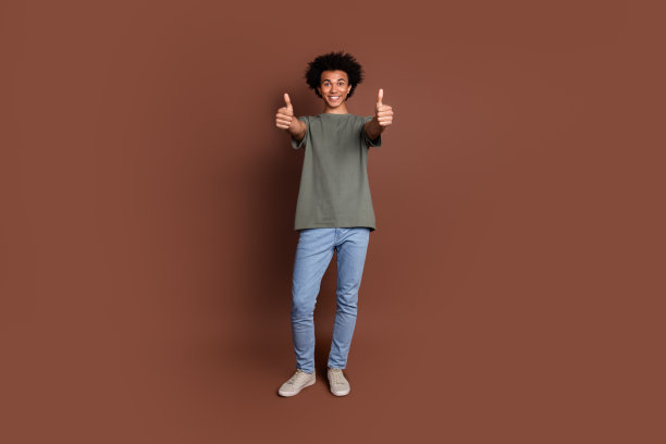 年轻人的全身长度的照片，波浪的头发在灰色t恤和牛仔裤给像反馈两个大拇指孤立的棕色背景图片下载