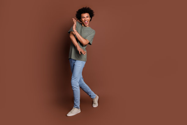 身穿灰色t恤和牛仔裤休闲服装的卷发青年在棕色背景上跳舞的全身照片图片下载