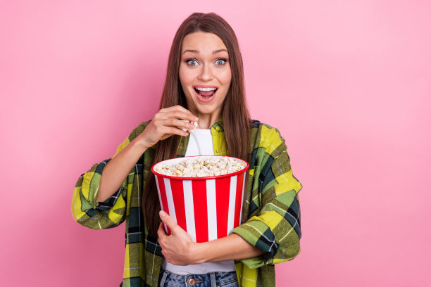 印象深刻的开朗女孩的照片，露齿微笑，张开嘴，拿着大爆米花盒子看电影，孤立的粉红色背景图片下载