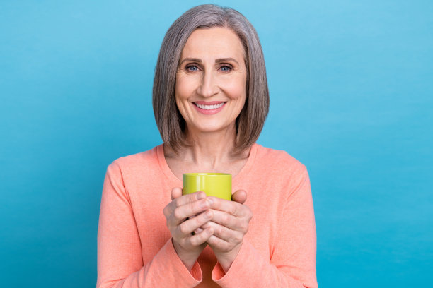 照片的好心情愉快的漂亮的祖母穿着粉红色的套头衫，拿着一杯咖啡享受早晨孤立的蓝色背景图片下载