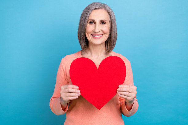 照片中风趣开朗的女子穿着珊瑚色衬衫，手拿红纸微笑，心隔蓝色背景图片下载