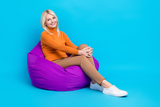 满意的漂亮女人的全尺寸照片橙色高领裤坐在豆袋咖啡休息隔离在蓝色背景图片下载