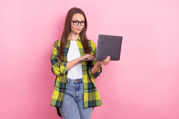 满意的可爱女人的照片，长发型，格子衬衫，看着笔记本电脑，阅读文档，孤立的粉红色背景图片下载