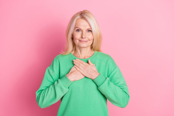 漂亮的退休妇女的照片肖像触摸胸部健康的心脏穿着时髦的绿色服装孤立在粉红色的背景图片下载
