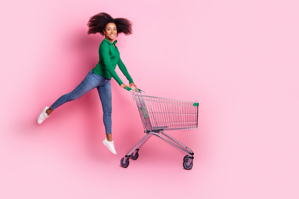 照片中有魅力的年轻女子推着超市购物车，穿着时髦的绿色衣服，在粉红色的背景上显得很有趣图片下载