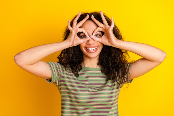 时髦的好心情女孩的照片，波浪状的头发，穿着条纹t恤，在黄色背景上显示双筒望远镜图片下载