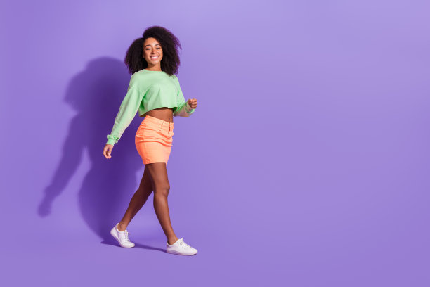 年轻自信的行走模特女孩的全身照片，波浪状的头发，绿色上衣和橙色迷你裙孤立在紫色背景上图片下载