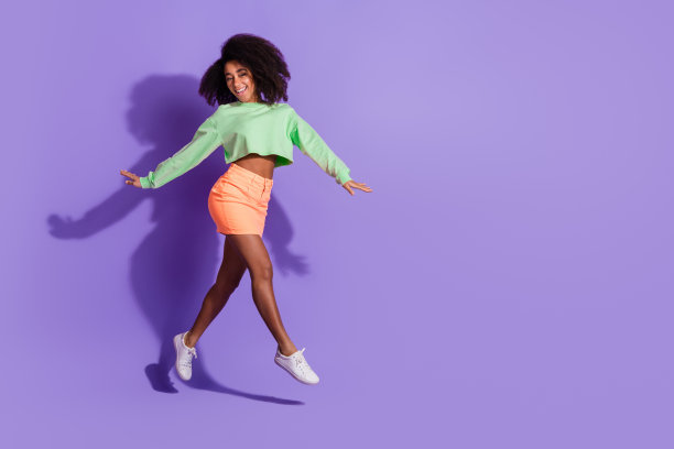 年轻有趣的蹦床女孩的全身长度的照片，波浪的头发在绿色的顶部和橙色的迷你裙孤立的紫色背景图片下载