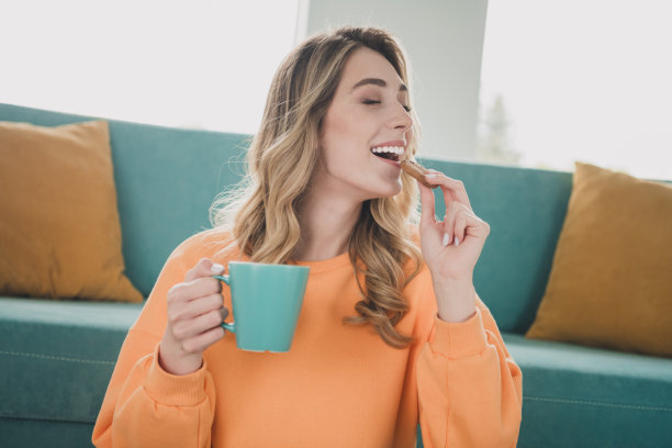 照片肖像迷人的年轻女子喝茶吃饼干穿着橙色的衣服舒适的日光家室内客厅图片下载