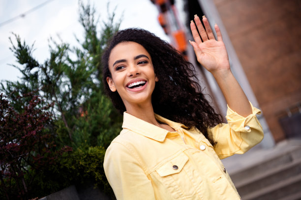 照片中，年轻的女孩面带微笑，留着波浪发型，穿着黄色时尚夹克，学生挥手问候，在户外度过城市周末图片下载