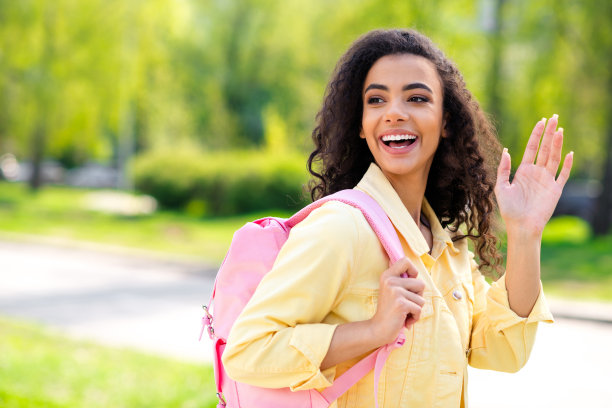 照片漂亮的卷发好心情女孩波浪手掌hi粉红色背包穿黄色夹克走在城市外的城市公园图片下载
