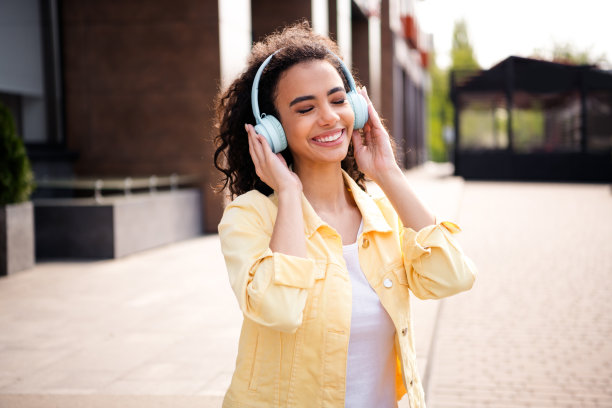 照片中，年轻的休闲女孩，卷发，穿着黄色时尚夹克，触摸着响亮的耳机，在户外度过周末图片下载