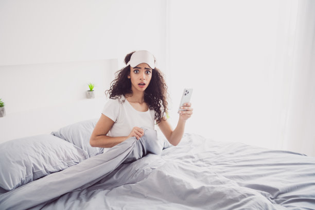 迷人的年轻女子的照片，坐在床上吓坏了，拿着装置，早上好，醒来的家卧室灯光白色的内部图片下载