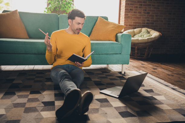 成熟男人的全尺寸照片坐在地板上在线学习上网本视频通话谈话穿着黄色的衣服舒适的日光家室内客厅图片下载