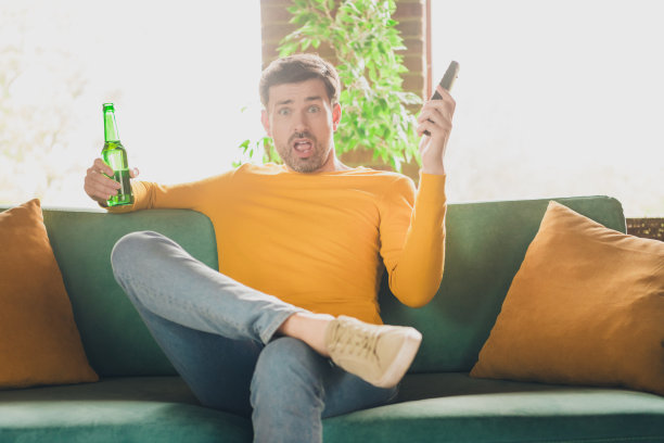 年轻人的照片肖像坐在沙发上看电视喝啤酒烦恼惊讶穿着黄色的衣服度过消遣宽敞的房子在室内房间图片下载