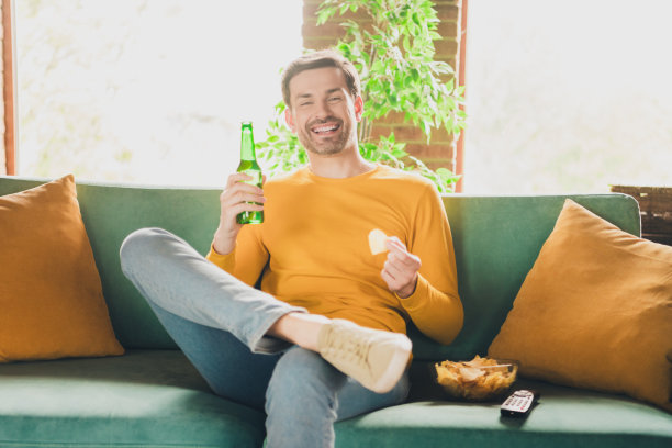 照片肖像英俊成熟的男人坐在沙发上看电视喝啤酒吃薯条穿着黄色的衣服舒适的日光家室内客厅图片下载